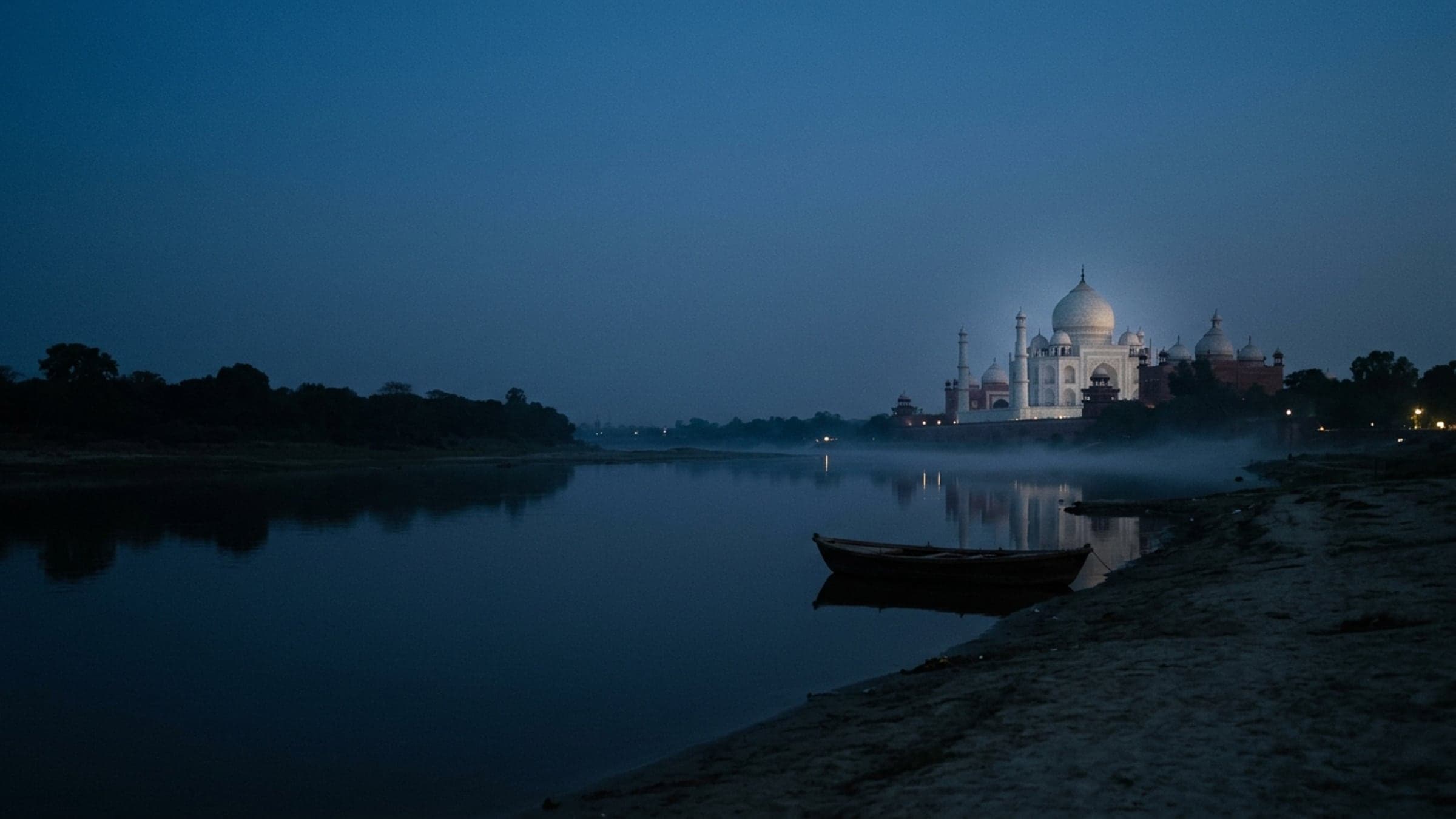 The Taj Mahal lit at blue hour, seen across the Yamuna with a wooden boat moored on the near bank